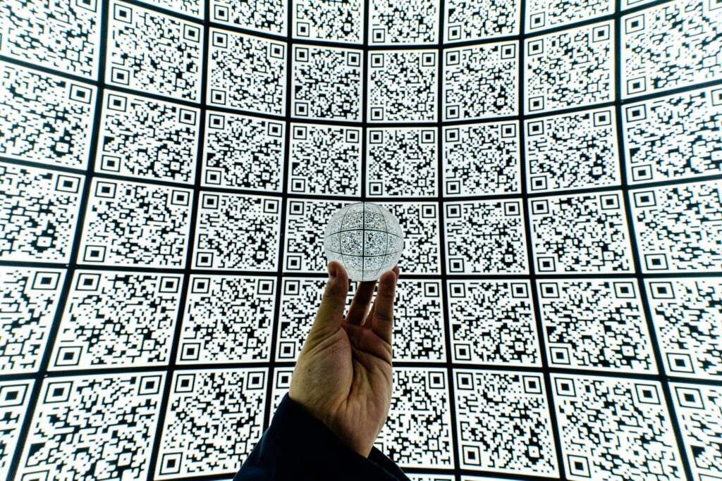 Hand holding a clear sphere with QR codes, against a curved wall of multiple QR codes.