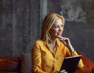 Smiling blonde woman in yellow shirt holding a tablet, sitting on a brown leather couch.