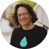 Smiling man with long dark hair and glasses wearing a black t-shirt with a light blue droplet design.