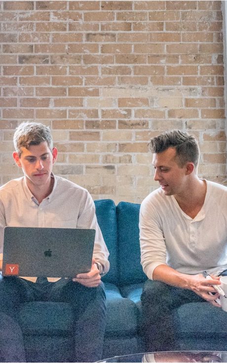 Two men on a blue couch, one looking at a laptop, the other watching him while holding a cup.