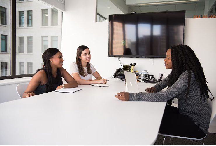 Three women in an office meeting, one speaking with a laptop, two listening and smiling.