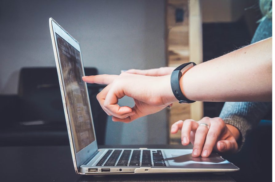 Two hands interacting with a laptop, one pointing at the screen and the other on the trackpad.