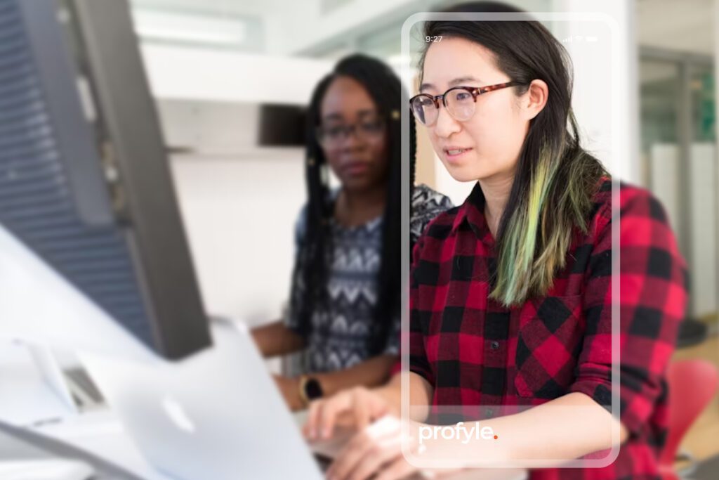 Woman in glasses and plaid shirt working on laptop, another person blurred behind her. 'profyle.' text in foreground.