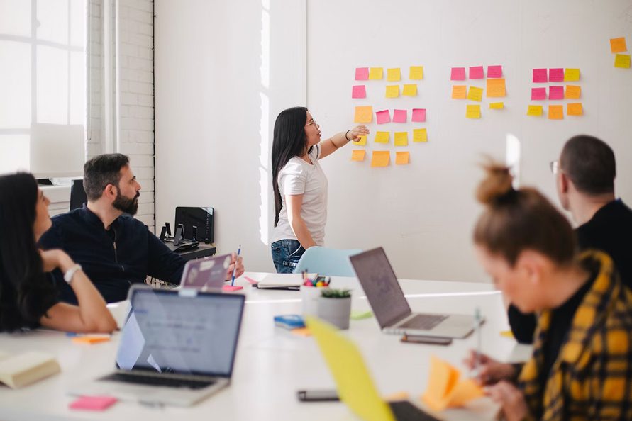 Woman points at sticky notes on a white wall during a meeting with colleagues at a table with laptops.