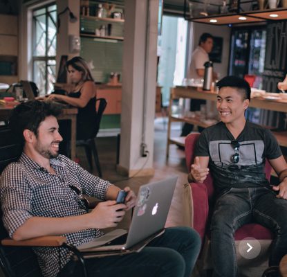 Two men smiling and talking, one with a laptop, in a busy indoor space with other people.