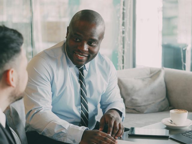 Two men in business attire discussing, with one smiling, at a table with coffee and a tablet.
